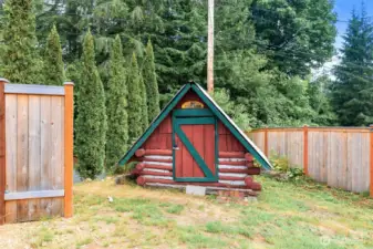 A whimsical, child-sized log cabin playhouse called “Kash’s Kabin,” complete with red logs, green trim, and an A-frame roof—adding personality and charm to the property.