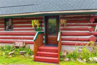Charming log cabin entry with a solid wood door, colorful hanging baskets, and rustic red-and-white tamarack logs, offering a warm welcome into the home.