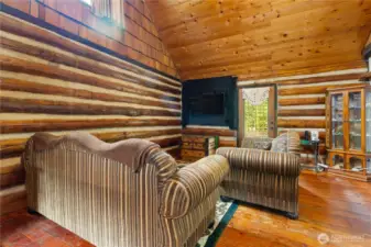 Spacious living room with exposed log walls and vaulted wood-paneled ceilings. A cozy sitting area is centered around the TV and antique-style furnishings, blending rustic comfort with cabin character.