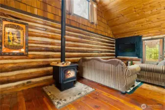 The living room showcases soaring pine ceilings and classic log walls, anchored by a cozy wood-burning stove set on a stone hearth. It’s the perfect place to curl up on a cool evening, with natural light streaming in and enough space for comfortable seating.