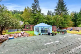 View of the spacious backyard featuring the large deck, a pop-up canopy for shade, and multiple rustic outbuildings. Surrounded by evergreens, this versatile outdoor space is perfect for gardening, gatherings, or creative projects.