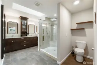 Spacious primary bath with dual-sink vanity, large walk-in shower with custom tile work, seating bench, and multiple showerheads including body sprays. Skylight and ample lighting make this space bright and inviting.