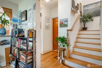 From the entry past living room on the right is the stairs going up to the primary bedroom and 2 others. Door in the background is the main floor bathroom down the hall left background goes to the kitchen and stairs going to the lower level. Left foreground leads to living room and kitchen.