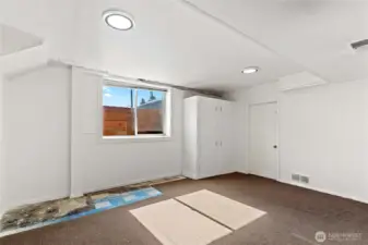 Bedroom with generous natural light from a large window, dark carpeting, and clean white walls. Includes a built-in cabinet for clothing and a discreet door housing the furnace and water tank, making it both functional and comfortable.
