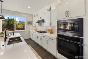 Kitchen  Full-height white cabinetry and quartz surfaces define this open concept kitchen, designed for modern Pacific Northwest living in Bothell’s Canyon Park area. A generous island with integrated sink anchors the space, complemented by a gas cooktop, wall oven, built-in microwave, and under-cabinet lighting. Expansive rear windows invite natural light while wide plank flooring adds warmth and contrast. The layout supports seamless flow between cooking and gathering, reflecting the elevated finishes expected in luxury new construction within the Northshore School District.    These photos are from a past Robinson Plan at a different community, so overall plan, colors, building orientation, finishes, home outlook, and other details can and WILL vary. All photos, renderings, site maps, and floorplans are for visualization purposes only.