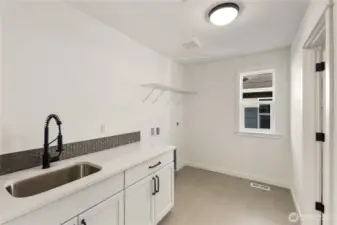 Laundry Room  This dedicated laundry room features quartz countertops, a utility sink with matte black fixture, and upper shelving for organized storage. Durable tile flooring and clean-lined cabinetry create a practical workspace designed for everyday efficiency. A window brings in natural light, enhancing both function and comfort.    Connected to the primary suite through the walk-in closet, the layout offers convenient direct laundry access while maintaining separation from main living areas. Located in Bothell near Canyon Park amenities, this thoughtfully placed space reflects the smart design found in contemporary new construction within the Northshore School District.    These photos are from a past Robinson Plan at a different community, so overall plan, colors, building orientation, finishes, home outlook, and other details can and WILL vary. All photos, renderings, site maps, and floorplans are for visualization purposes only.