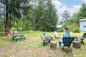 Cleared yard bordered by evergreens and featuring a rustic fire pit area with Adirondack chairs and picnic table—ready for s’mores, stargazing, and storytelling.