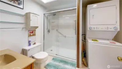 Well-appointed primary bath featuring a spacious step-in shower with glass enclosure and grab bars for added comfort. Includes built-in shelving, overhead skylight for natural light, and convenient in-unit laundry with a stacked washer and dryer.