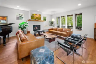 The main-floor living room is anchored by a striking white tile fireplace with a wood mantle. Hardwood floors and large windows to an outdoor deck fill the space with warmth and natural light.