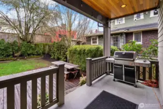 The covered rear deck with wood-planked ceiling steps down to a fenced backyard with lawn, mature plantings, and a lower patio area — a practical outdoor setup for grilling and gathering year-round.