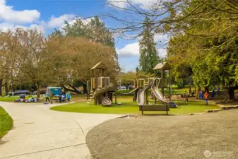 The park's nature-inspired play area features dual playground structures for different age groups, set among mature trees with plenty of lawn space for kids to roam.