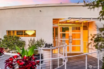 A dramatic entry sets the stage — the curved approach and glass double doors hint at the light-filled elegance within, as a sparkling chandelier glows like a beacon of luxury