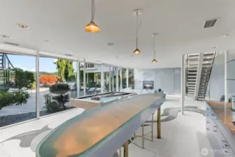 The recreation level features a sleek glass-topped bar cocktail table, a pool table, and walls of windows that open to the patio and waterfront.
