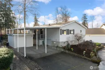 Charming front exterior showcasing a spacious covered carport, attached garage, and elevated front porch. Mature trees and landscaped garden beds create a peaceful and inviting first impression.