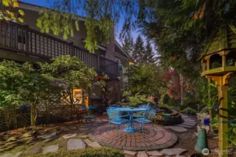 Lush landscaping surrounds the brick patio, blending vibrant greenery with colorful planters. A serene, fenced setting ideal for morning coffee or a relaxing evening.