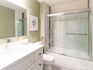 Primary bathroom boasts large vanity, newer toilet, newer tub with tiled tub surround, newer shower doors, and extra nook with storage rack for towels or reimagine the space with your very own personal touch!