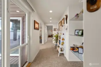 Wide, bright hallway, illuminated by natural light, features built-in glass shelving for displaying art or collectibles, enhancing the open and airy feel. Leading into a spacious primary suite, media/game room, and provides easy access to the hot tub deck, seamlessly connecting key living areas with style and functionality.