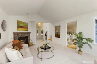 This cozy living room offers a welcoming transition to the rest of the home, complete with vaulted ceilings, a gas fireplace, and soft natural light. A peek into the entryway and kitchen reveals the home’s open, connected layout.
