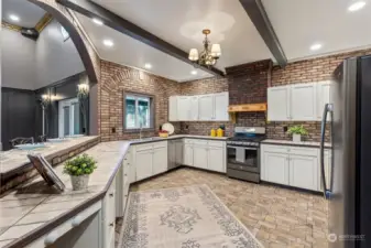 Classic white Shaker cabinetry has beautiful rustic brick as a backdrop, tile floors below and beam accentuated ceilings with enough counterspace to encourage multiple cooks to collaborate!