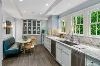 With a full wall of divided-light windows ushering in garden green, the Kitchen balances precision and polish. Waterfall-edge marble countertops and a custom glass hutch underscore both function and beauty—an inviting space where mornings begin with light and leisure.