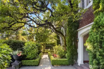 The path to the Main Gate unfolds beneath the sweeping branches of a heritage oak—one of many layered elements in the home’s tapestry of landscape and legacy. With clipped boxwood, ivy-clad brick, and dappled sunlight, this garden passageway feels both stately and alive.