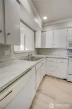 Entrance to kitchen from Living room. Updated countertops with grey veining, shaker-style upper & lower cabinetry with silver knob finishes, and a double basin stainless kitchen sink. Dishwasher located to left of sink near one entrance to kitchen. Sink sits under the pass-through window. Recessed light installed throughout the ceiling for a clean look. NEW LVP floor with a light wood color.