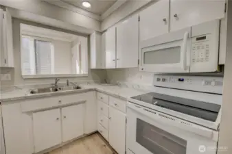 Refreshed kitchen with new LVP flooring and updated countertops with grey veining. Shaker-style cabinets with silver knob finishes.  White electric glass stove stop with over-the-range matching white microwave. Pass-through window looking out to window bringing extra light into the kitchen. Laundry room situated in on the other side of kitchen where pass-thru leads to.