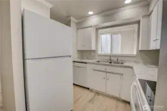 L-shaped kitchen with two entrances. Entrance pictured is from hallway. Features white appliances, updated LVP flooring and new countertops with grey veining. Handle-free Refrigerator with top freezer; glass cook top on the stove/range.