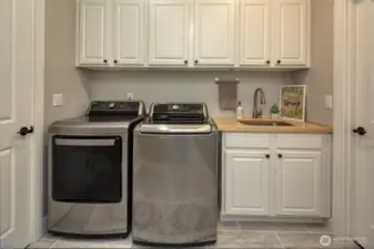 Crisp and functional, this laundry room offers sleek stainless steel appliances, ample upper and lower cabinetry, a butcher block countertop with deep utility sink, and stylish tile flooring—designed for both practicality and charm.