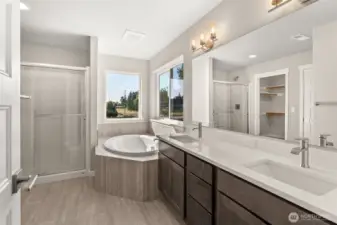 Five-piece primary bath features dual vanity with quartz counters, tile flooring, and tile deck around deep, soaking tub. Walk-in shower features a bench and sliding glass door with brushed nickel finish.