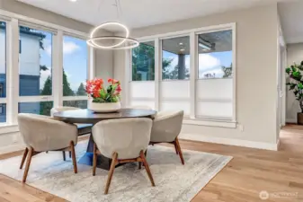 A light-filled dining area wrapped in nearly floor-to-ceiling windows, bringing in soft natural light and glimpses of the surrounding evergreens. The sculptural modern chandelier anchors the space, creating an elegant setting for everyday dinners or gatherings with friends.