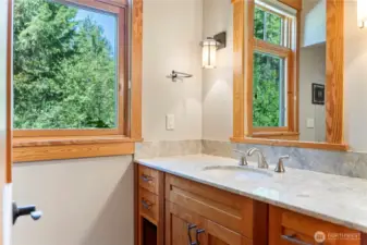 Peaceful main floor bath with natural stone counters, custom wood cabinetry, and windows that frame the lush forested setting.