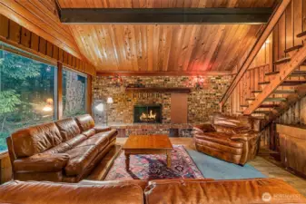 The living room feels plucked from a woodland tale—brick fireplace standing tall, beams overhead, and windows that frame the trees like living art. Sunlight shifts with the hours, casting a glow that makes even the stairs look adventurous, winding you up toward rooms tucked under the rafters.