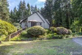 The north side of the house has a grassy area for parking, with a stairway up to the deck and pathway to the front door. Private and peaceful, and a gardener's paradise. The window in the photo is the breakfast nook in the kitchen.