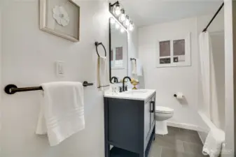 Spa-like full bath completely remodeled with a sleek charcoal vanity, matte-black fixtures, modern globe vanity lighting, white subway-tiled tub/shower combo, large window for natural light, and luxury vinyl plank flooring. Clean, contemporary, and magazine-ready.