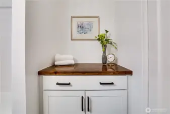 Handsome custom-built storage piece with warm butcher-block top, two soft-close drawers, and double-door cabinet below. Crisp white shaker doors with matte-black pulls add extra style and function to this fully remodeled bathroom – perfect for towels, toiletries, and everyday essentials.