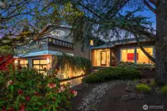 This captivating Northwest contemporary home features warm cedar siding and a welcoming entrance framed by lush, mature landscaping. The twilight view showcases the home's elegant lines, with a balcony overlooking the beautifully maintained front yard. Illuminated windows reveal glimpses of a bright, stylish interior with modern accents. The thoughtful landscaping includes vibrant rhododendrons, ornamental trees, and a stone drainage path that enhances both beauty and functionality. This property combines Pacific Northwest charm with sophisticated design, creating an inviting sanctuary nestled within a tranquil garden setting.