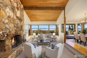 The living room features vaulted ceilings, a striking floor-to-ceiling wood-burning fireplace, and a wall of west-facing windows framing breathtaking Sound and mountain views.