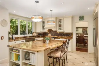 This inviting kitchen features rich natural stone countertops, a hammered copper farmhouse sink, and creamy shaker-style cabinetry. A mosaic tile backsplash adds texture behind the stainless-steel range, while pendant lights warm the central island. Large windows overlook lush greenery, and the open layout flows into a cozy living area with a stone fireplace—perfect for everyday living and entertaining.