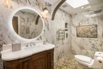 Step into opulence in this artfully designed bathroom featuring dramatic marble walls, a custom wood vanity with integrated LED mirror, a curbless walk-in shower with mosaic flooring, rainfall showerhead, towel warmer, and an elegant in-shower tapestry-style tile mural that brings timeless sophistication to every detail.
