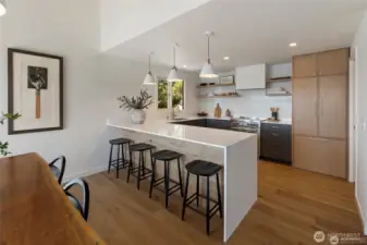 This spacious, light-filled kitchen blends luxury and function with an oversized peninsula, sleek open shelving, premium appliances, and seamless flow to the dining area and outdoor deck.