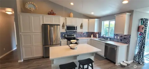 Bright and beautifully appointed, this modern kitchen blends coastal style with everyday ease—featuring white cabinetry, gleaming stainless appliances, quartz countertops and an elegant blue and white glass backsplash. A central island with pendant lighting and seating invites connection, all framed by warm wood floors and natural light. Effortlessly stylish, perfectly Surfside.