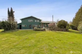 View of the back side of home from the grassy yard, mature landscaping and terraced garden beds lead to the patios. The yard is partially fenced. There is a spacious tool & wood shed as well as a shop that's approximately 75'x25'. Just under an acre.