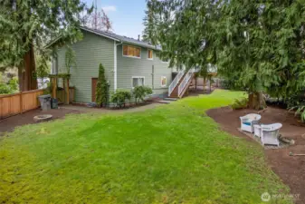 A wide look at the level lawn and outdoor seating area, with towering evergreens that enhance privacy and shade. This yard offers a great blend of open space and natural landscaping.
