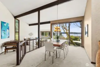 This is the moment. You walk in and that wall of windows completely takes over, framing the water like artwork. Just outside, a moss-covered mature tree rises up in its own alcove between the house and deck, creating this layered, almost curated natural feature that makes the view feel even more special. The space itself leans into its 1973 roots in the best way, with the dining area slightly elevated beside the living room, giving it that classic split-level vibe that feels both retro and refined. Vaulted ceilings with dark exposed beams add contrast and structure, while the mix of cream carpeting, warm dining room tones, and crisp white in the living room keeps everything bright and balanced. It’s a little dramatic, a little sophisticated, and completely memorable.