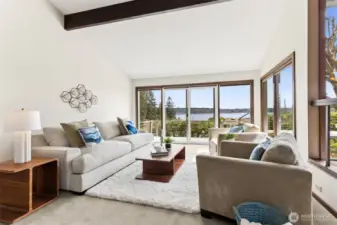 All eyes go straight to the windows, and for good reason. The living room is framed by a full wall of glass that captures the water view from nearly every angle, with a slider that makes stepping outside feel effortless. The vaulted ceiling and exposed beam give the space a little architectural edge, balanced by soft flooring and clean white walls that keep things light. Outside, the deck’s cable-style railing disappears into the background, letting the view take over without interruption.