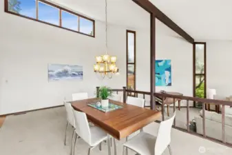From the dining area, you get a full appreciation for the layered design that makes this home stand out. A few steps down lead into the living room, creating that classic 1970s split-level feel that adds dimension without closing anything off. The vintage chandelier anchors the dining space with warmth and character, while high above, a long panoramic window at the peak of the vault pulls in sky views and soft natural light. As your eye moves downward, those tall, narrow accent windows in the living room stretch from floor to ceiling, bringing in even more light and that unmistakable architectural style of the era. A dark wood railing frames the edge of the dining area, adding both safety and definition while still keeping everything open and connected.