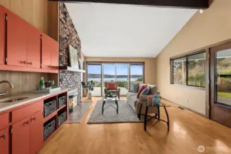 The view pulls you in immediately, straight through the family room and out to Budd Inlet. From the kitchen vantage point, the connection between spaces feels easy and natural, perfect for staying part of the conversation without missing a beat. Vaulted ceilings open everything up, while the hardwood floors keep it warm and grounded. The original brick fireplace holds onto that 1970s character, now paired with a gas insert so it actually gets used. Light reflects beautifully off the white ceilings and plays against the soft suede-toned walls. Off to the side, the doorway leads to a covered patio, giving you another way to step into that waterfront setting without even trying.