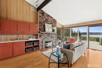 There’s a lot happening here, and it all works together. The fireplace brings in texture and warmth with its multitone brick, while the family room’s built-in beverage station adds a layer of convenience you don’t always see. With its prep sink, storage above, and open shelving below, it creates a natural spot for drinks, coffee, or casual hosting. It feels intentional, not like an afterthought. Across the room, the full wall of windows frames the water view in a way that’s hard to compete with, and the sliding doors lead right out to the deck, extending the space even further.