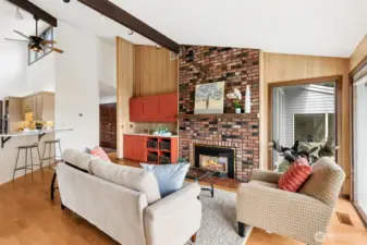 From this spot near the window, the fireplace really takes center stage, stretching all the way from floor to vaulted ceiling and giving the room a strong vertical moment. The exposed beams overhead add that architectural detail you don’t see every day, and the vertical panel accent wall brings in warmth that balances out the scale of the space. Off to one side, the built-in beverage station adds function right where you want it, with its own sink and storage creating a natural spot for drinks and hosting. Then your eye moves down the line and you catch the connection to the rest of the home, a glimpse of the front entry and the bar-height counter seating that opens into the kitchen. It’s a layout that keeps everything connected without feeling crowded, ideal for both everyday living and entertaining.